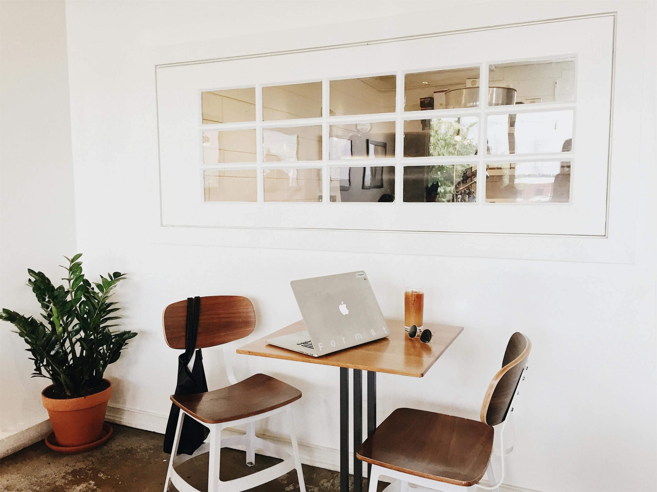 A modern, minimalist workspace featuring a laptop on a table, wooden chairs, and a potted plant.